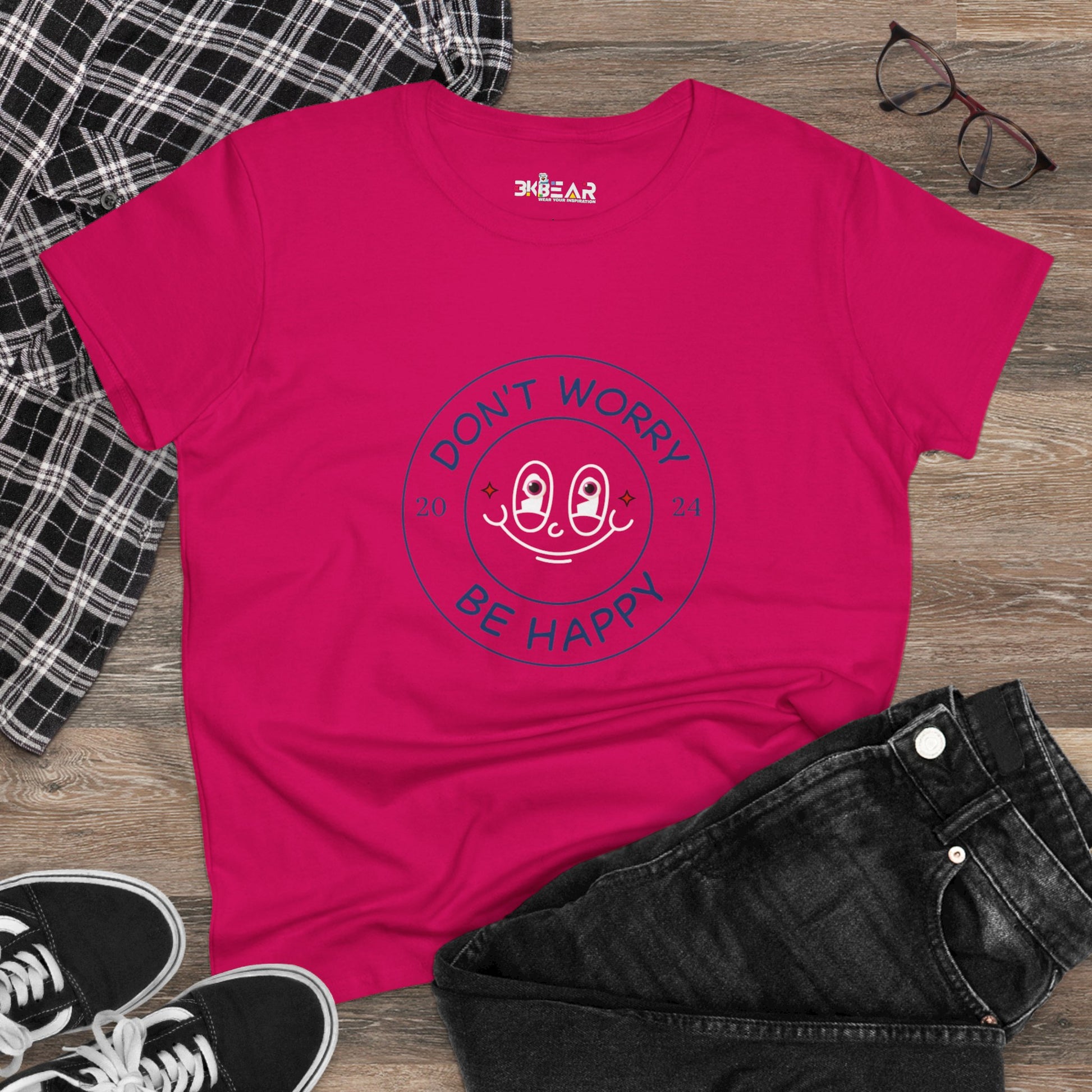 Bright pink don’t worry be happy t-shirt with a smiling face design laid out with jeans, sneakers, and glasses