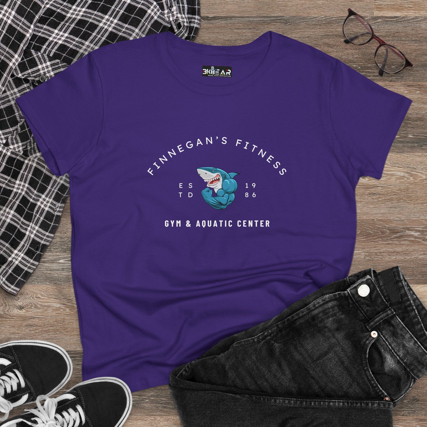 Purple workout shirt women graphic with Finnegan’s Fitness shark design and gym text laid on wooden floor