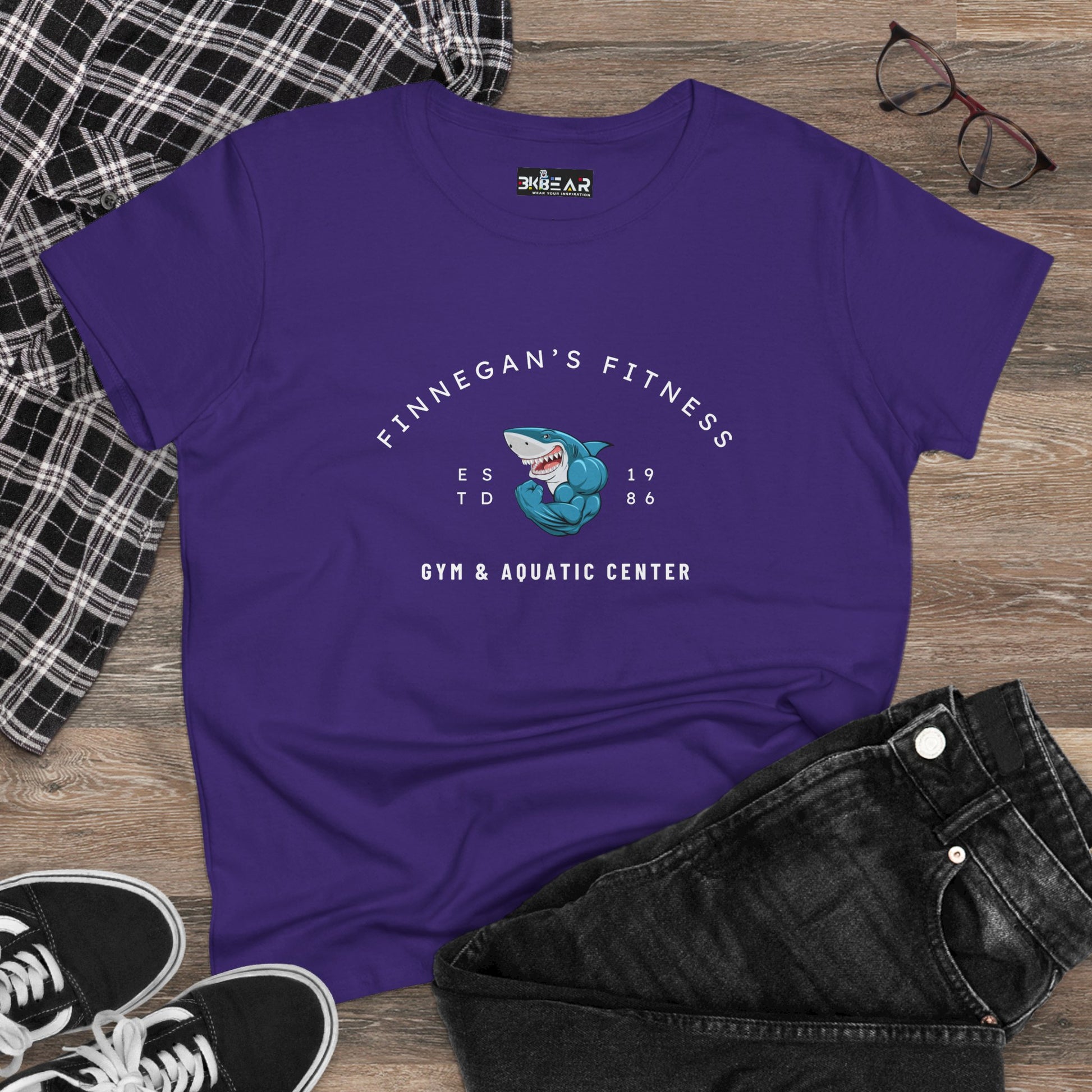 Purple workout shirt women graphic with Finnegan’s Fitness shark design and gym text laid on wooden floor